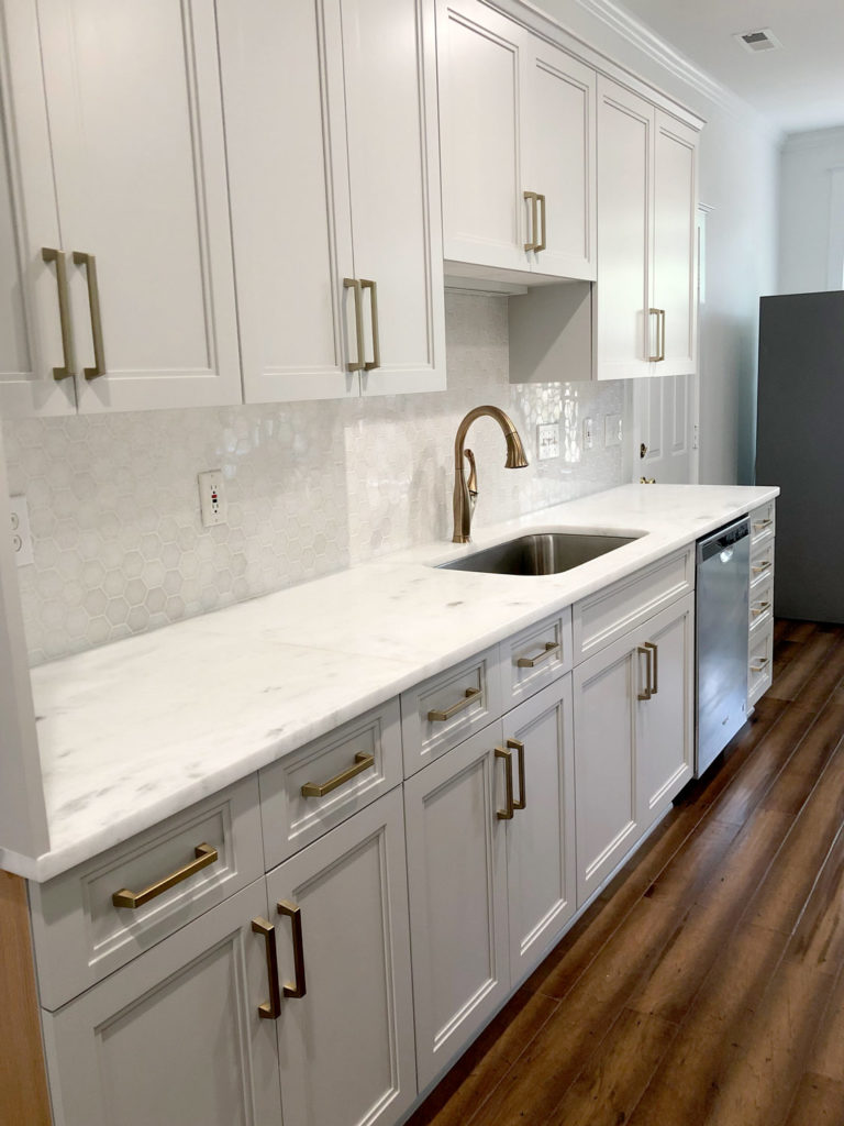 Refaced white kitchen cabinets with white tile backsplash. A marble countertop with brass curved faucet and stainless sieel sink.